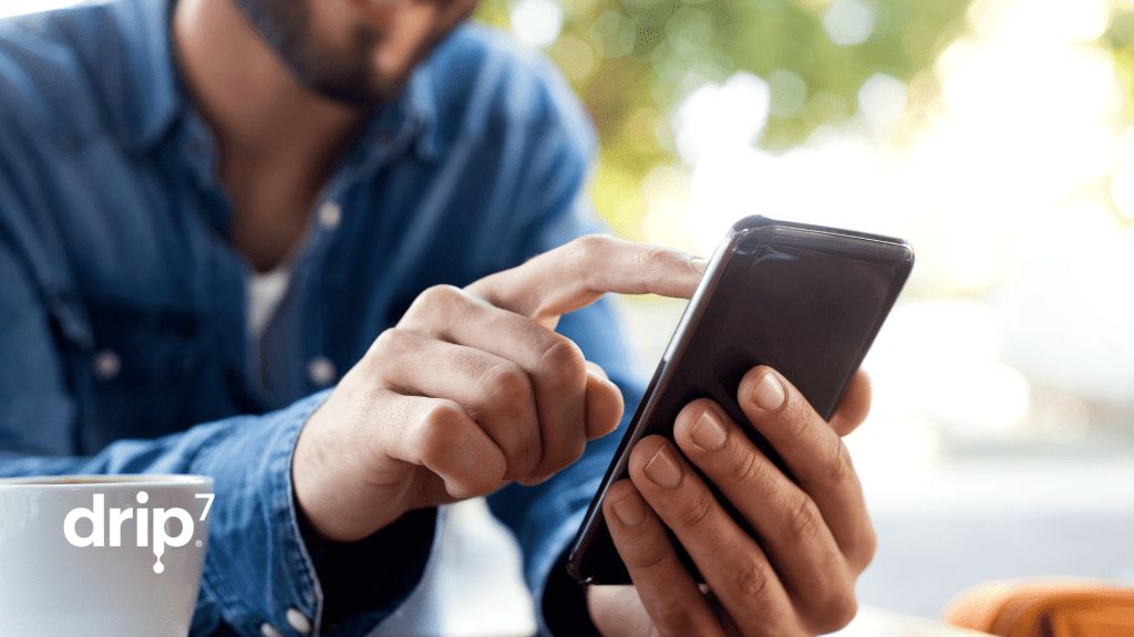 Man types on cell phone in a coffee shop, possibly doing Drip7 cybersecurity training